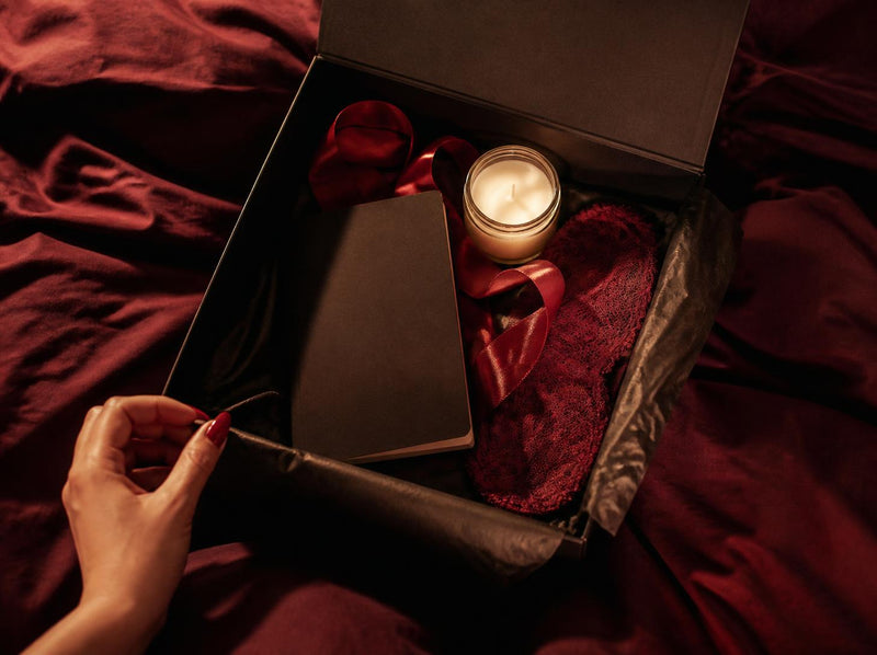 Between the Sheets spicy romance box opened on wine satin with a candle glowing inside